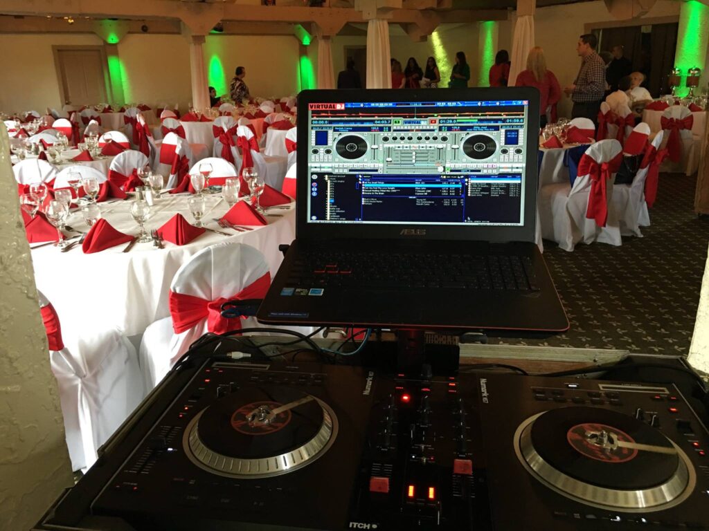 DJ turntables and laptop in front of a wedding reception with red and white table settings and green uplighting